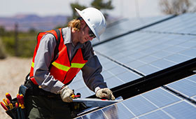 Junger Mann mit Helm und orangener Warnweste arbeitet an einem Solarpanel auf einem Hausdach